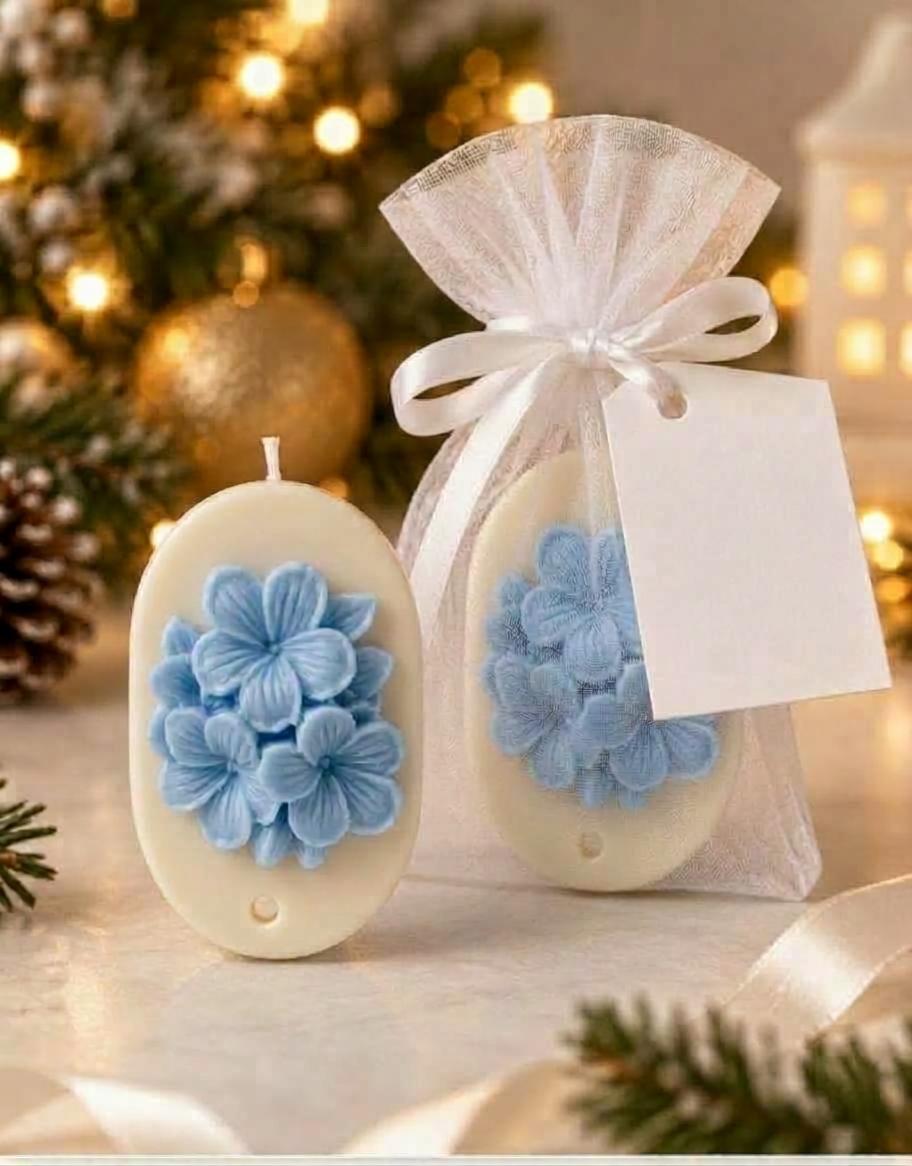 Vela artesanal oval branca com flores azuis em relevo, apresentada em saquinho de organza branco com laço de cetim